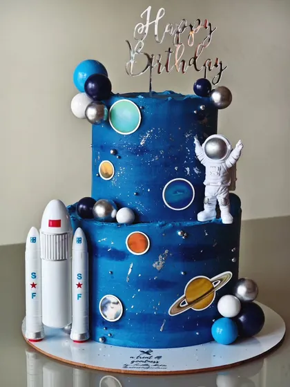 An out-of-this-world space-themed cake for a young astronaut's birthday. This two-tiered buttercream cake captures the galaxy with a deep blue palette, silver leaf accents, and fun elements like planets, rockets, and an astronaut topper.
