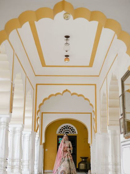 A beautiful bridal portrait in a palace corridor. The repeating arches create a stunning frame for the bride in her elaborate pink and multi-colored lehenga, showcasing her elegance.