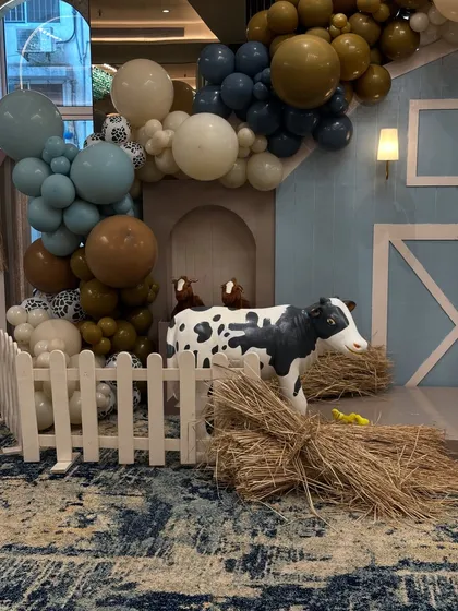 A close-up of the farm setup, with a white picket fence, hay bales, and a friendly cow prop, all framed by a beautiful balloon arch.
