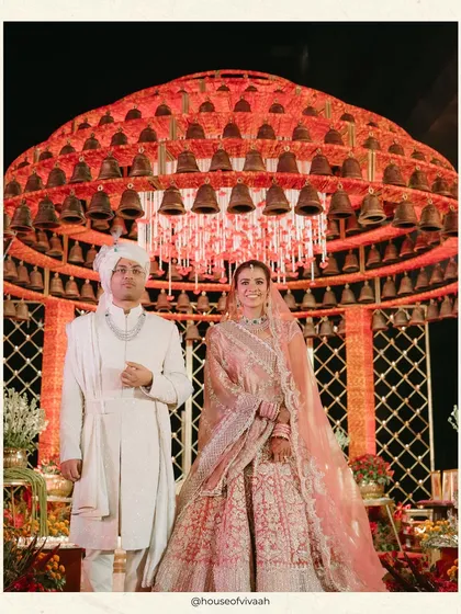 The couple stands before the magnificent bell mandap, a structure designed to symbolize blessings and divine harmony. The scale and detail of the installation create a powerful and unforgettable wedding portrait.