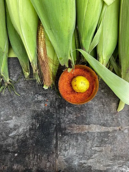 A simple, beautiful flatlay of fresh corn, with a lemon and a bowl of red chilli powder ready for seasoning.