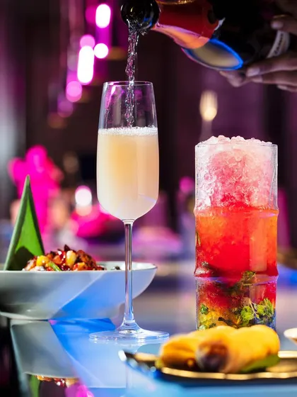 This shot captures the vibrant, neon-lit ambiance of Hakkasan. A champagne pour into a flute is juxtaposed with a colorful crushed-ice cocktail, reflecting the restaurant's energetic and modern setting.