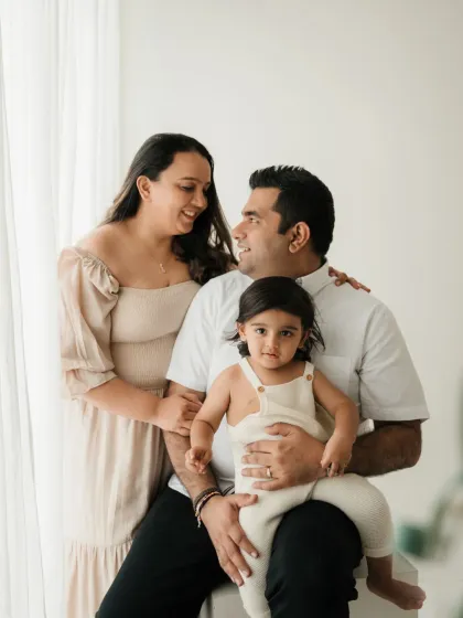 A relaxed family portrait where the parents look at each other while their child sits on dad's lap. This captures a natural, unposed moment.