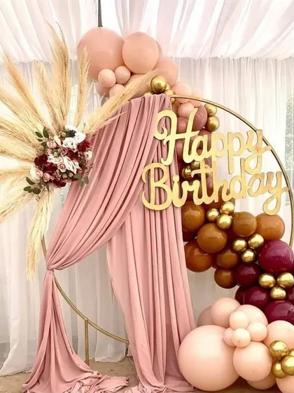 A chic and rustic birthday decor concept, combining a gold arch with pampas grass, drapery, and a modern balloon garland.