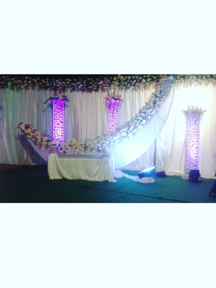 This stage design features a graceful arc of white flowers against a simple draped backdrop. Illuminated pillars add a touch of grandeur, making it a versatile option for receptions, haldi, or mehendi events.