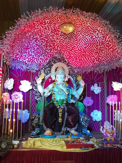 This large-scale Ganpati pandal showcases a creative use of materials, with a backdrop made of pink and white checkered patterns and large, handcrafted paper flowers. The result is a festive and visually striking stage for Bappa.