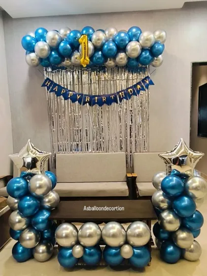 A clean and stylish first birthday decoration in blue and silver. The setup includes a balloon garland over a silver curtain backdrop, flanked by two balloon pillars topped with star-shaped foils.