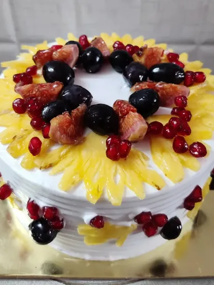 A delightful fresh fruit cake topped with pineapple, figs, grapes, and pomegranate seeds arranged in a beautiful pattern.