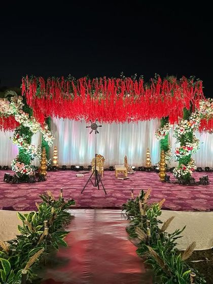 A grand and dramatic reception stage dominated by a ceiling of hanging red florals. The stage is framed by lush green and white floral pillars, creating a bold and passionate statement.