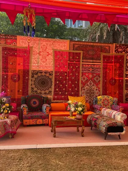 Another seating arrangement at the Kathiyawadi-themed event, with a backdrop of layered red and orange patterned rugs and textiles. The design creates a warm, inviting, and culturally authentic atmosphere.