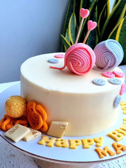 A special cake for an 80th birthday, themed around knitting and Indian sweets. It features fondant yarn balls, buttons, and miniature sculpted sweets like jalebi and ladoo.
