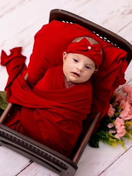 Wrapped in a vibrant red swaddle, this baby rests in a tiny wooden bed. The bold color contrasts beautifully with the light wood floor, making for a striking and memorable newborn picture.