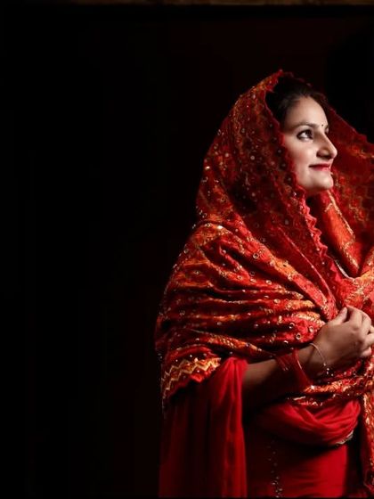 A beautiful portrait of a woman in a traditional red Punjabi phulkari dupatta. The dramatic lighting highlights the intricate embroidery and her joyful expression during the ring ceremony festivities.