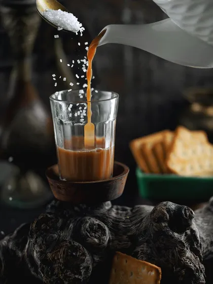 A classic Indian chai scene with a dynamic pour and sugar sprinkle. The dark, rustic styling and the traditional cutting glass evoke a sense of authentic, street-style comfort.