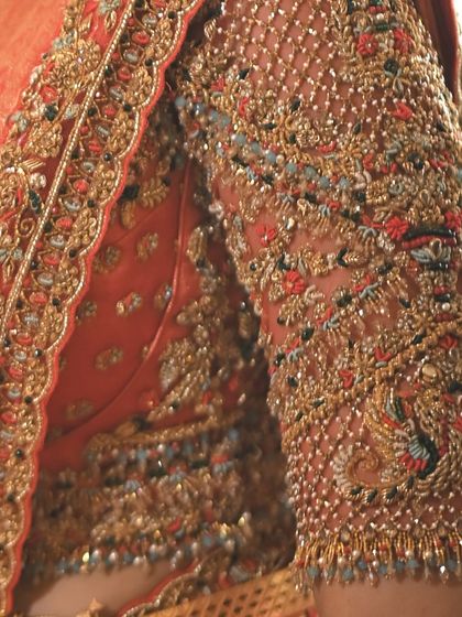 An extreme close up of an embroidered sleeve on an orange silk blouse. You can see the texture of the Zari thread, the shimmer of the beads, and the precision of each stitch.