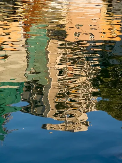 The reflection of colorful buildings ripples on the surface of the water. This abstract phone photograph plays with color, light, and texture to create a painterly effect.