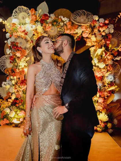 The couple shares an intimate moment against a stunning photo backdrop made of woven fans and an arch of autumn-toned florals, showcasing a unique and artistic installation.