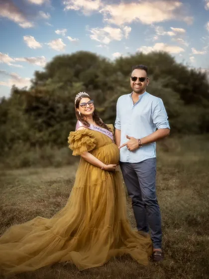 A fun and candid outdoor shot. The dad-to-be proudly points to the baby bump, and her joyful laugh is infectious. It’s a happy, relaxed moment captured under a beautiful sky.