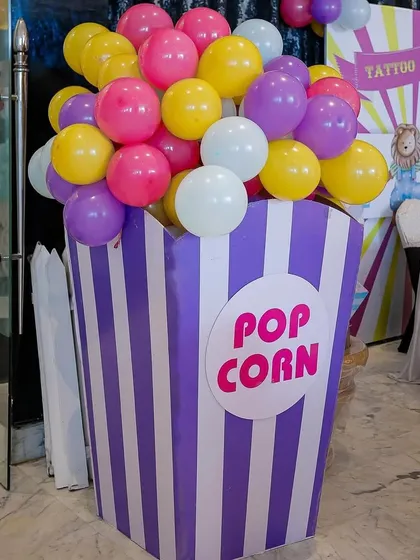 A giant popcorn prop filled with bright balloons adds a fun, oversized element to this carnival party's decor, seen here in a purple and white striped design.