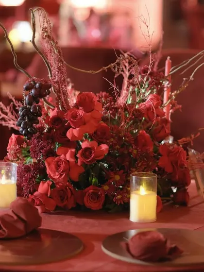 A dramatic, monochromatic red tablescape for a reception dinner. The centerpiece combines red roses, dried branches, and grapes, all bathed in the warm glow of candlelight for a passionate and intimate atmosphere.