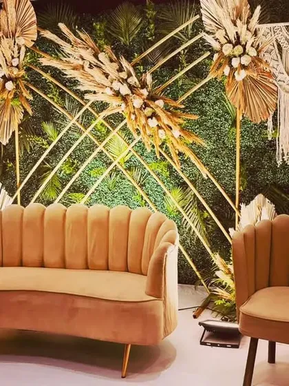 A chic boho-themed stage with a green foliage wall, gold geometric frames, and macrame hangings. Dried pampas grass and palm leaves complete this earthy and stylish look, perfect for a modern, nature-loving couple.