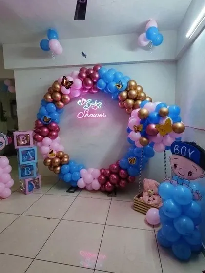 A circular balloon ring is a stylish alternative to a traditional arch for a baby shower or gender reveal. This design uses a mix of pink, blue, and gold balloons, B-A-B-Y blocks, and character cutouts, all centered around a glowing "Baby Shower" neon sign.