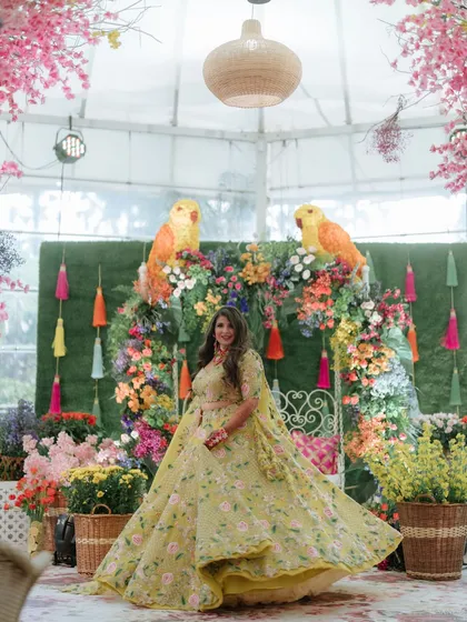 A bride twirls in her vibrant lehenga against a backdrop of colorful florals and parrot motifs, perfectly capturing the joyful spirit of a Mehendi function.