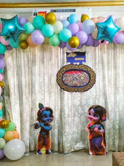 A naming ceremony setup at home with a pastel-colored balloon arch. The theme includes Radha Krishna cutouts and transparent 'BABY' blocks.