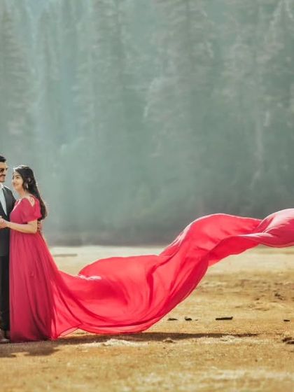 A beautiful, wide shot showing the couple in a vast, misty landscape. The vibrant pinkish-red gown provides a stunning focal point in the natural setting.
