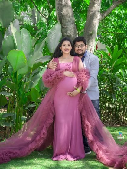 A full-length portrait of a couple in a lush garden. The long, flowing train of the maternity gown adds a touch of elegance and drama to this romantic outdoor scene.