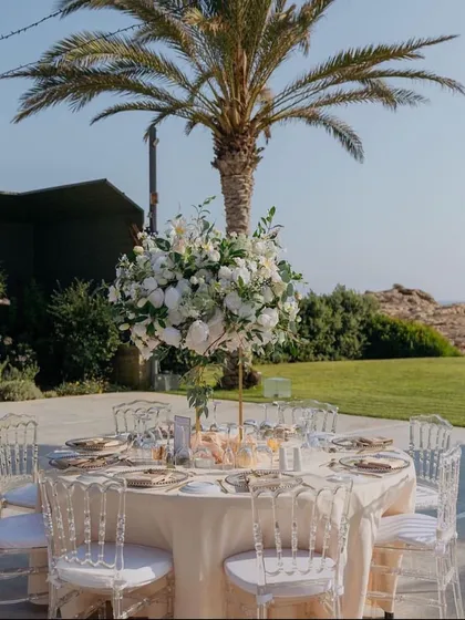 A classic round table setup for an outdoor wedding, with a tall floral centerpiece and clear Chiavari chairs, perfect for a sunny day.