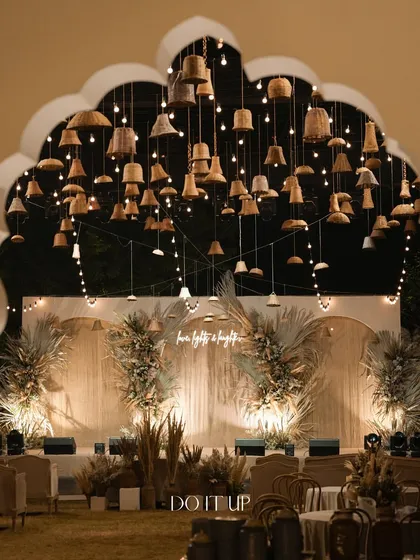 The main stage of the bohemian Sangeet. The backdrop features natural textures and dried palms, with an installation of hanging wicker lampshades and string lights creating a warm, magical canopy.