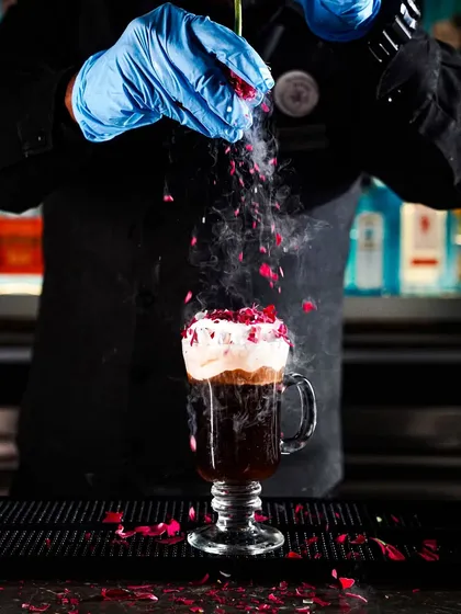 A bartender in blue gloves sprinkles dried rose petals over a steaming Irish coffee cocktail. This action shot captures the theatricality and craft of expert mixology.