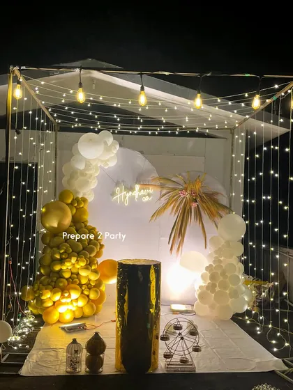 A versatile cabana-style backdrop illuminated with fairy lights and Edison bulbs. Decorated with gold and white balloon garlands and boho elements like dried palm fronds, this setup is suitable for birthdays, anniversaries, or even a bachelorette party.