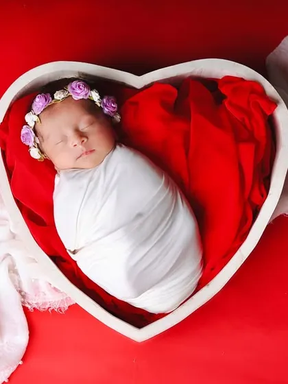 A bold and beautiful portrait. The baby is wrapped in clean white fabric, resting inside a heart-shaped bowl against a vibrant red backdrop, creating a striking symbol of love.
