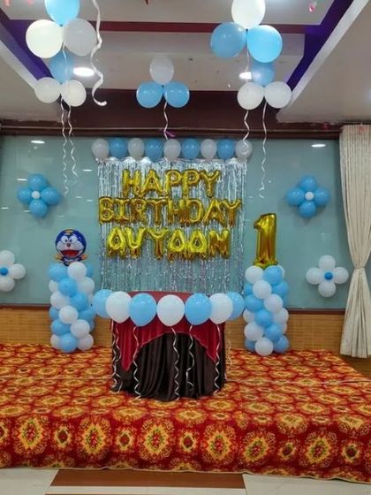 A classic birthday stage decoration with a silver foil curtain backdrop. The setup includes "HAPPY BIRTHDAY" foil letters, balloon pillars, and a number "1" foil balloon.