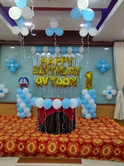 A classic birthday stage decoration with a silver foil curtain backdrop. The setup includes "HAPPY BIRTHDAY" foil letters, balloon pillars, and a number "1" foil balloon.