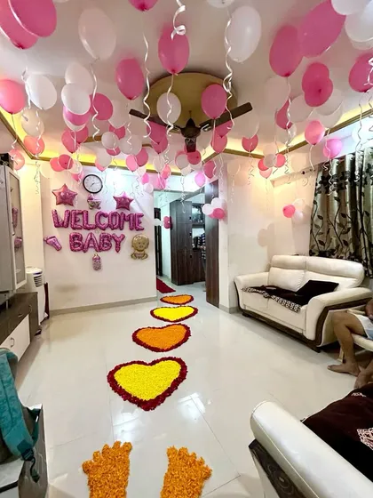 A beautiful and simple way to welcome a newborn home. This living room decoration includes pink and white balloons on the ceiling, "Welcome Baby" foil letters, and a heartfelt pathway of marigold flower hearts.