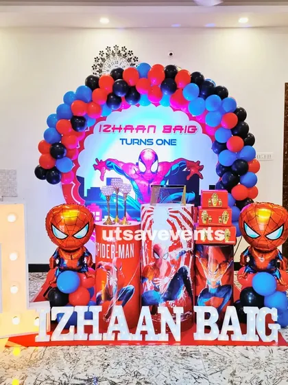 A Spiderman-themed first birthday party. The setup includes a character backdrop, Spiderman balloon figures, a light-up number '1', and themed plinths.