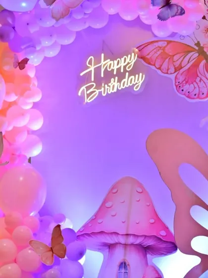 A close-up of the butterfly-themed birthday backdrop with a 'Happy Birthday' neon sign. The purple and pink lighting creates an enchanted feel.