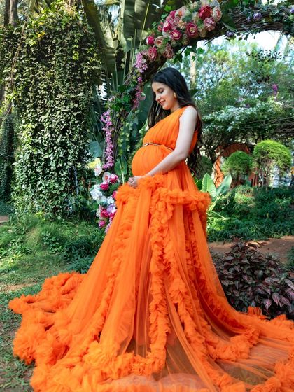 A stunning full-length shot of a mom-to-be in a vibrant orange gown, set against a backdrop of lush greenery and a floral arch.