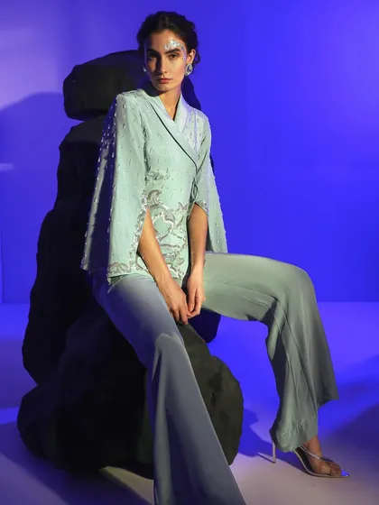 A model seated on the rock props, wearing a mint green embellished pantsuit. The pose is relaxed but powerful, showcasing a modern take on occasion wear.