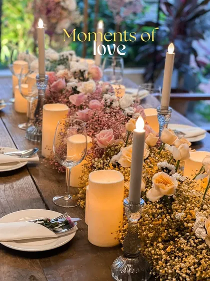 A close-up of a romantic tablescape, perfect for an anniversary dinner. The table runner is a lush arrangement of pink baby's breath and roses, interspersed with the warm glow of pillar candles.