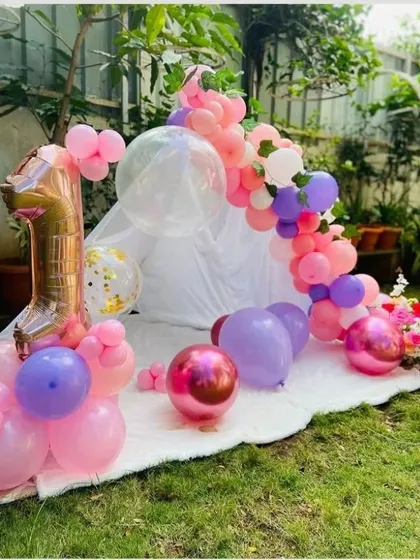 A cute and simple first birthday picnic setup in a garden. A small white tent is decorated with a garland of pink, purple, and chrome balloons, perfect for a cozy celebration.