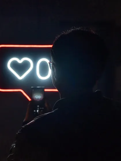 A visitor captures the neon installation on their phone, becoming part of the artwork's critique of digital life. The reflection in the phone screen adds another layer to the experience.