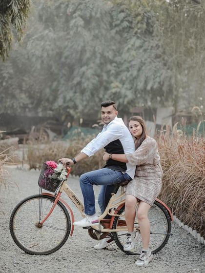A couple poses with a vintage bicycle on a gravel path, showcasing a classic and charming pre-wedding photo idea. The natural surroundings add to the rustic feel.
