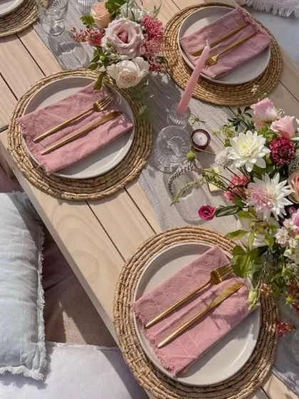 A closer look at a rustic-chic picnic table setting. The combination of natural wood, woven placemats, and soft pink linens creates a relaxed yet elegant feel.