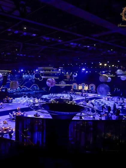 A wide shot capturing the full expanse of the cosmic-themed engagement party. The central bar, guest tables, and massive stage are all united under a deep blue, star-filled atmosphere.