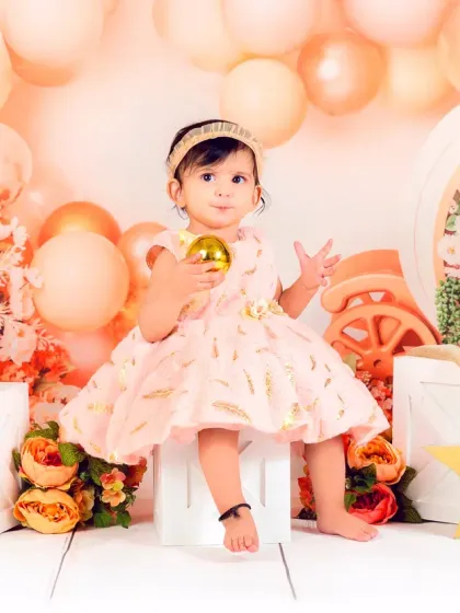 A baby girl sits elegantly in a peach-toned floral setup, holding a golden prop. She looks like a little queen on her throne.