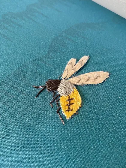 A tiny, delicately embroidered bee from our Folly design. This small detail is a testament to the thoughtful artistry and narrative depth we infuse into our wallcoverings, celebrating nature's subtle wonders.